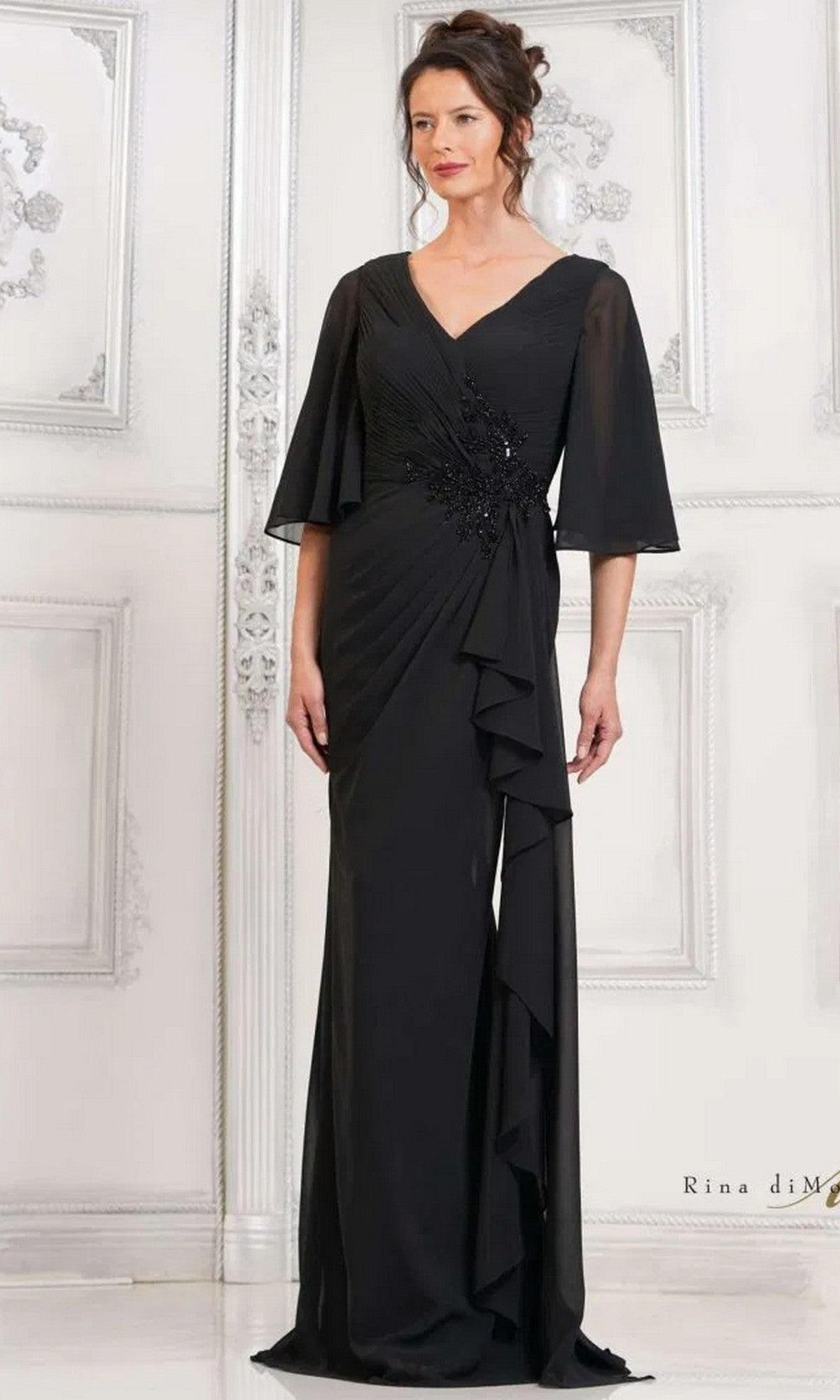 A woman stands before an ornate white wall wearing the Rina Di Montella RD2935 long black formal dress featuring chiffon fabric, a ruched bodice, flared sleeves, and decorative detailing.